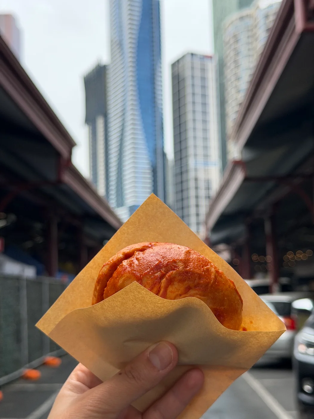 A hand holds a saucy SOSMOS burger between the market sheds with Melbourne towers behind